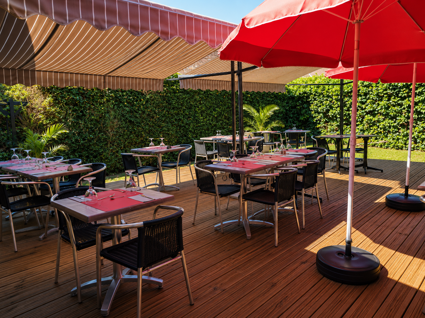 La terrasse d'O'trèfois : tables dressées sous les parasols rouges, entourées d'un mur de verdure.
